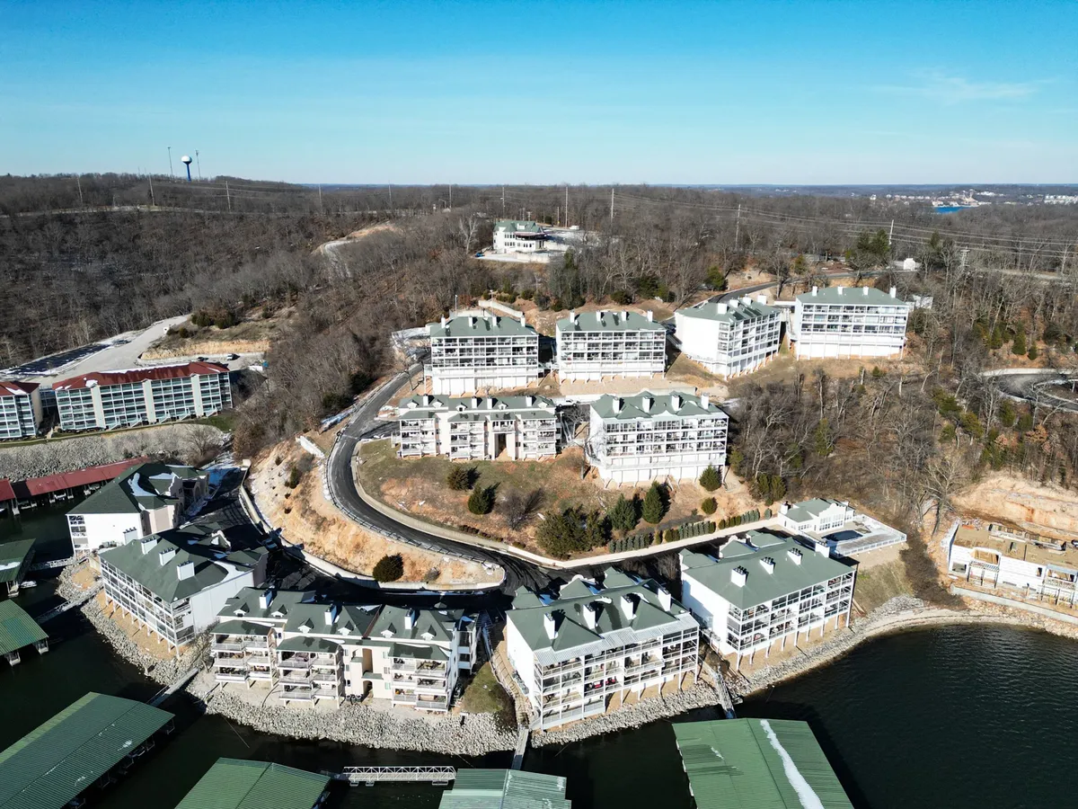 A broad exterior shot featuring the condominium buildings nestled by the waterfront, surrounded by trees and open spaces. The scene evokes tranquility while highlighting both the proximity to water for fishing and boating, as well as easy access to outdoor exploration.