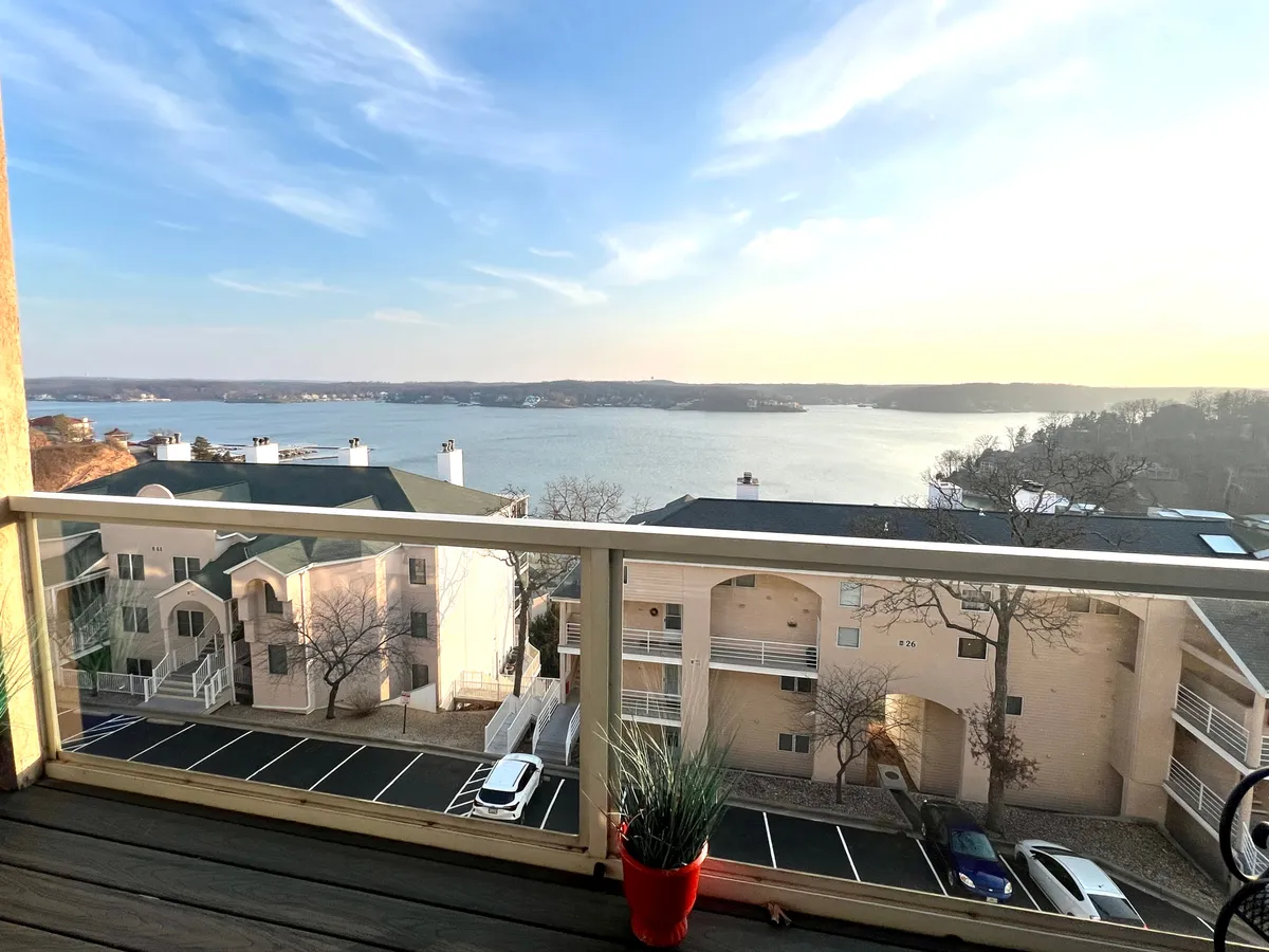 This image captures a serene lake view from a balcony, framed by a railing and a potted plant. The vantage point suggests a peaceful spot to start the day, take in the sunrise, or unwind after outdoor adventures.