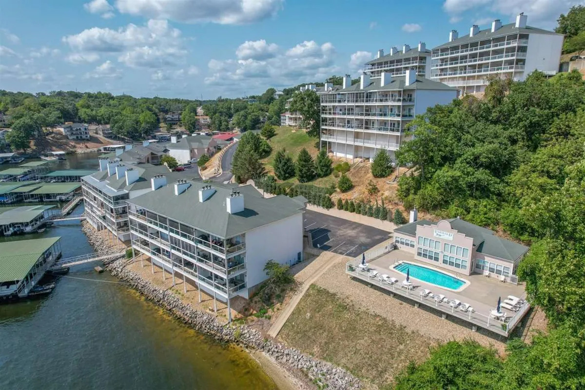 An expansive exterior shot featuring a gated community swimming pool with clear water, surrounded by lounge chairs and lush greenery. In the background, residential condo buildings overlook a tranquil lake vista, hinting at direct pool and water access. The scene evokes a sense of relaxation and opportunity for recreation, with potential for socializing poolside or enjoying serene moments by the water.