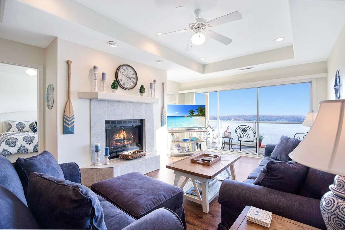 A cozy living room with a fireplace as the focal point, large windows framing sweeping lake views, and a plush seating arrangement ideal for relaxing or hosting gatherings. Natural light fills the space, and the inviting layout encourages both relaxation and socializing.