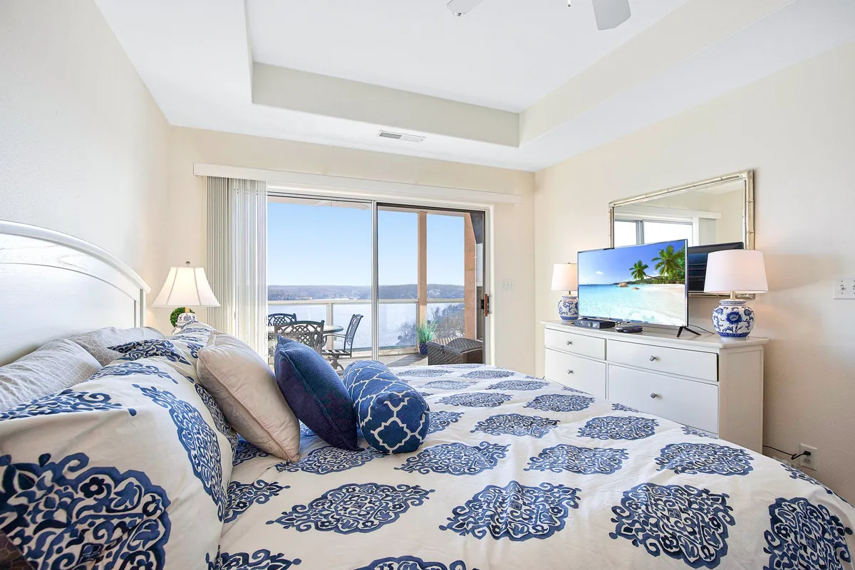 A tranquil bedroom featuring a bed with patterned bedding, a large window offering a beautiful lake view, a TV displaying a serene beach scene, and decorative lamps that add warmth to the space. The room is bathed in natural light, enhancing its restful atmosphere.