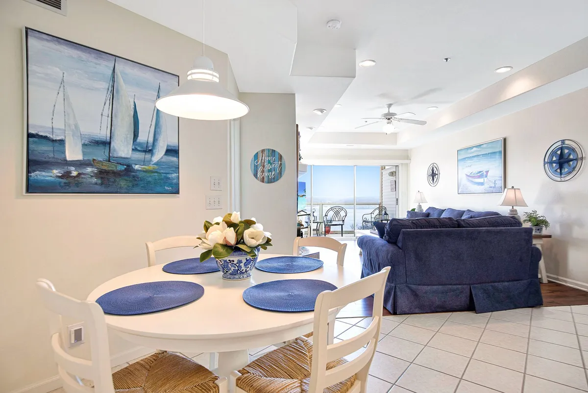 A cozy dining area with a dining table adorned with a floral arrangement, nautical artwork on the wall, and a comfortable blue couch nearby. The space is welcoming and arranged for shared meals or casual gatherings, with a subtle view suggesting proximity to water.