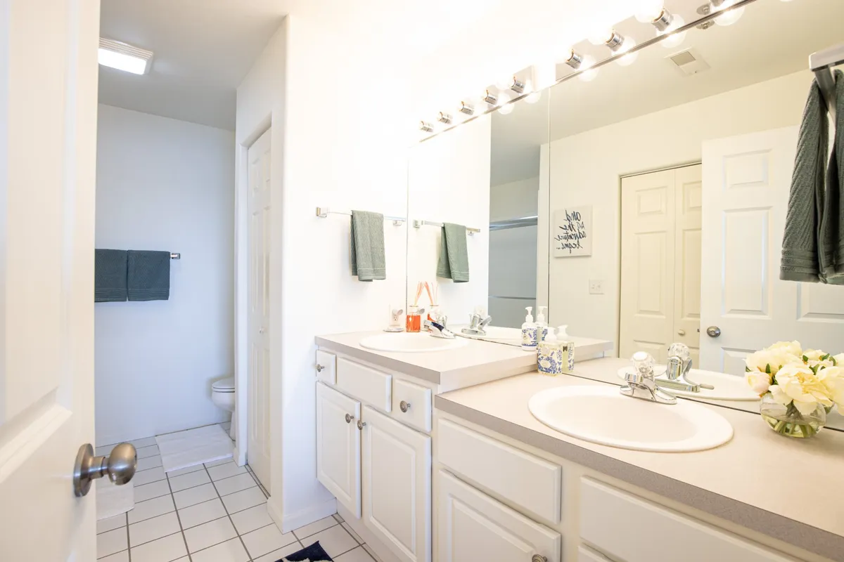A spacious, inviting bathroom with a double sink vanity, large mirror, floral arrangement, and plush towels. The space is bright and thoughtfully designed for daily routines and relaxation.